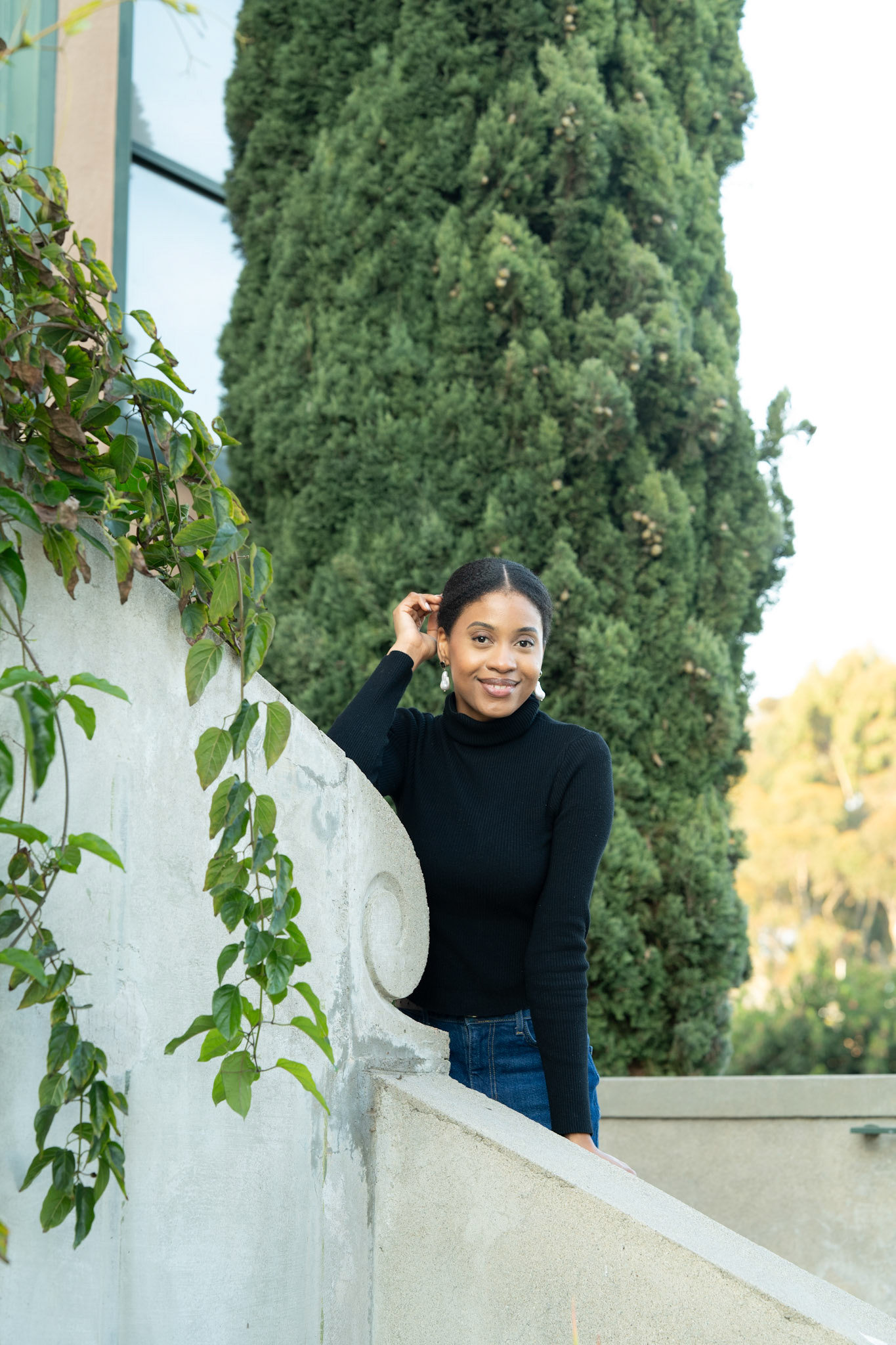 A smiling woman in a black turtleneck leans against a stone wall with greenery, set against a tall tree
